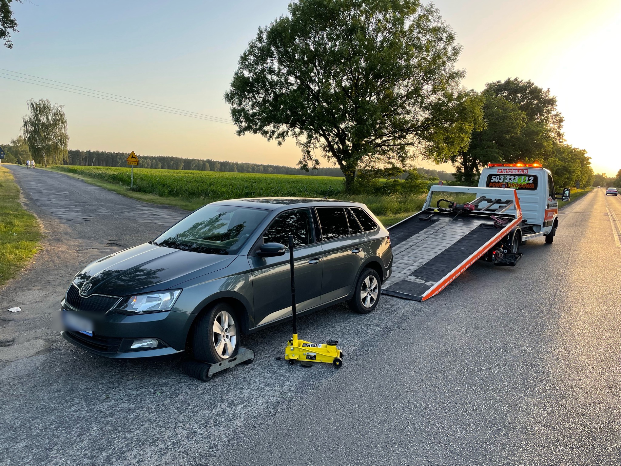 Szary samochód osobowy z uszkodzonym kołem, podnośnik hydrauliczny, laweta pomocy drogowej na drodze wiejskiej o zachodzie słońca.