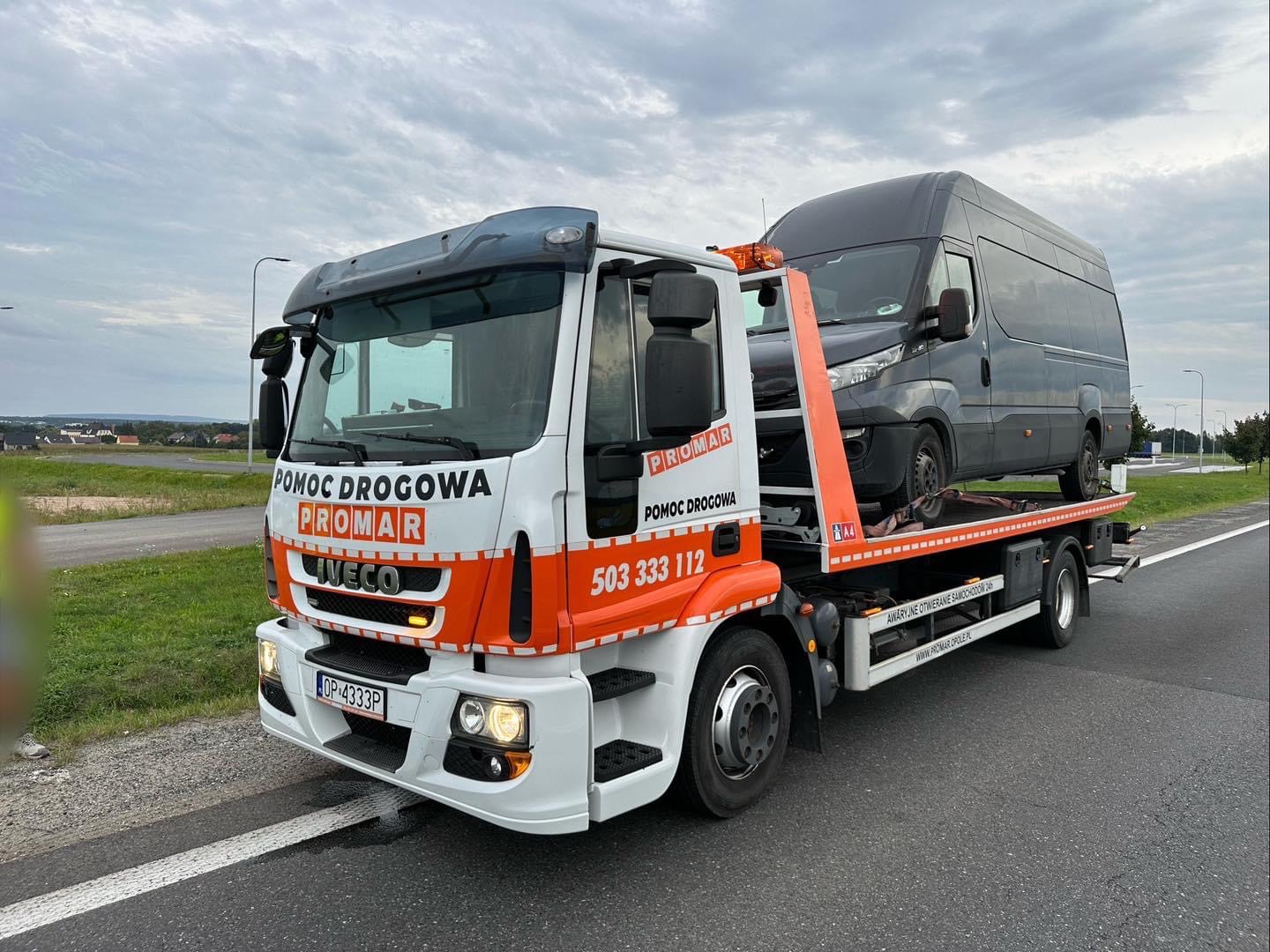 Laweta firmy PROMAR z Iveco na platformie, transportująca szary samochód dostawczy na tle pochmurnego nieba.
