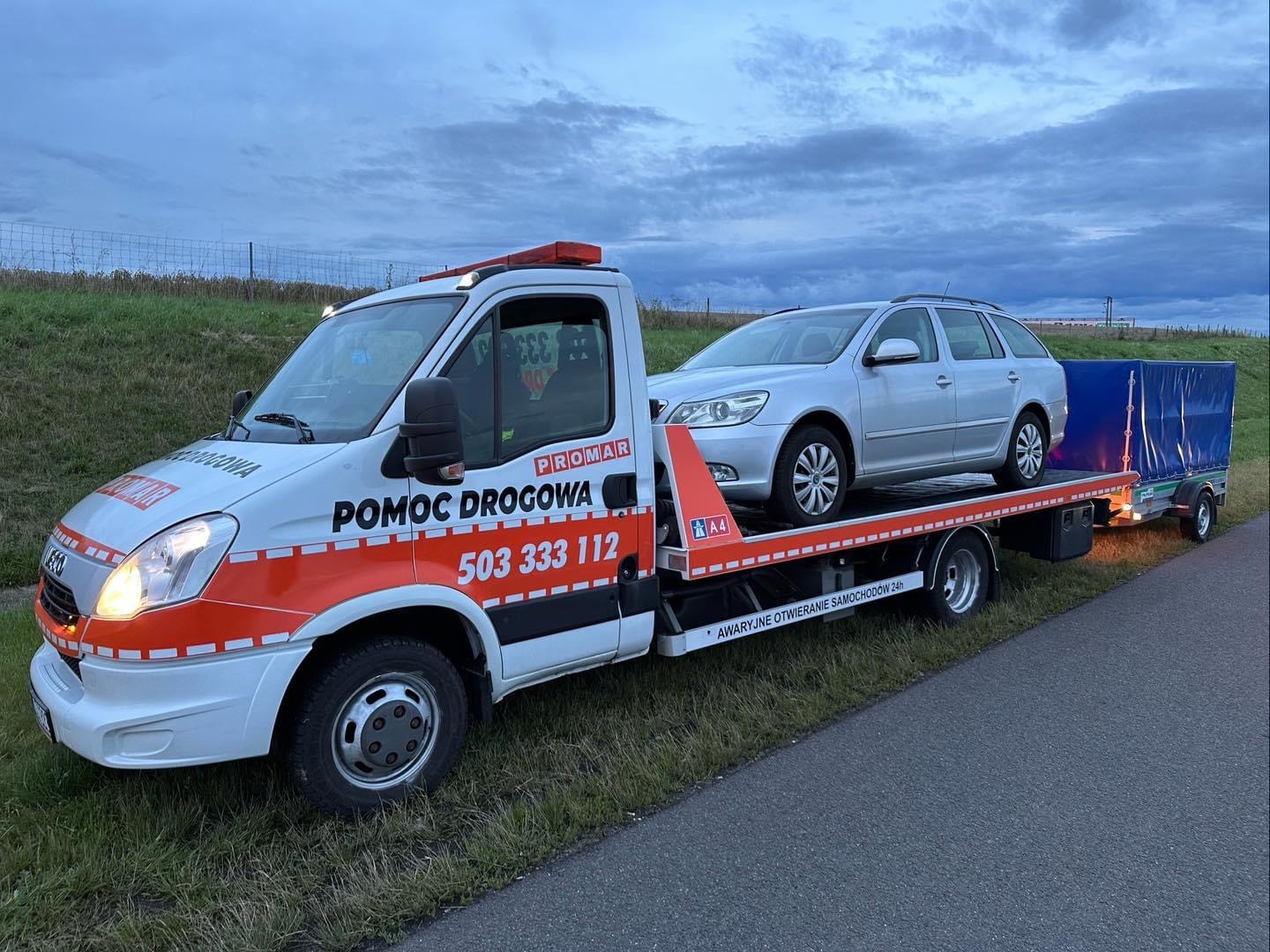 Laweta z załadowanym srebrnym kombi Skoda Octavia i niebieską przyczepką na tle zachmurzonego nieba i zielonego pola.