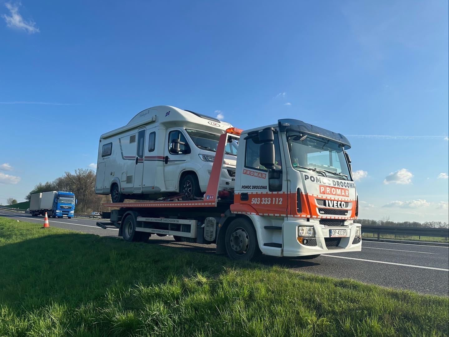 Laweta pomocy drogowej marki Iveco z pomarańczowymi elementami, przewożąca biały kamper na autostradzie w słoneczny dzień, widoczny numer telefonu 503 333 112 i logo firmy Promar.