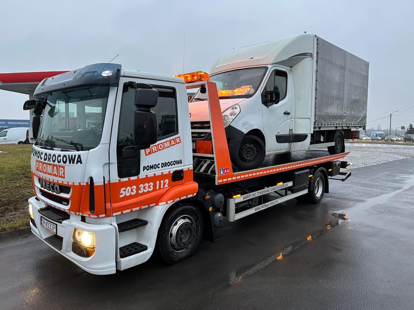 Laweta z pomarańczowym wysięgnikiem przewozi biały samochód dostawczy z plandeką na mokrej nawierzchni, w tle stacja benzynowa i ruch uliczny.