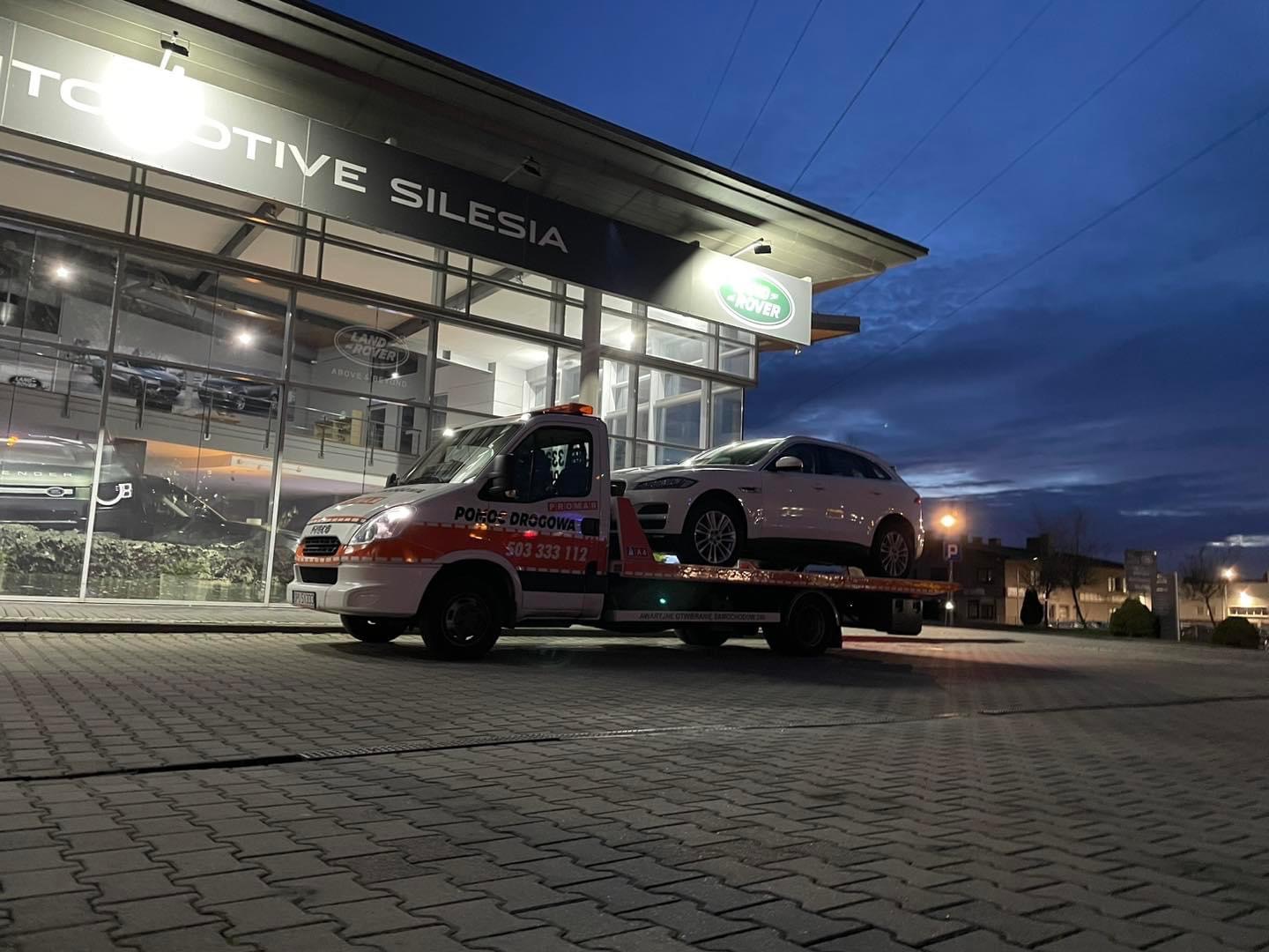 Laweta z białym SUV-em Jaguar na tle salonu samochodowego Land Rover/Jaguar Automotive Silesia o zmierzchu.