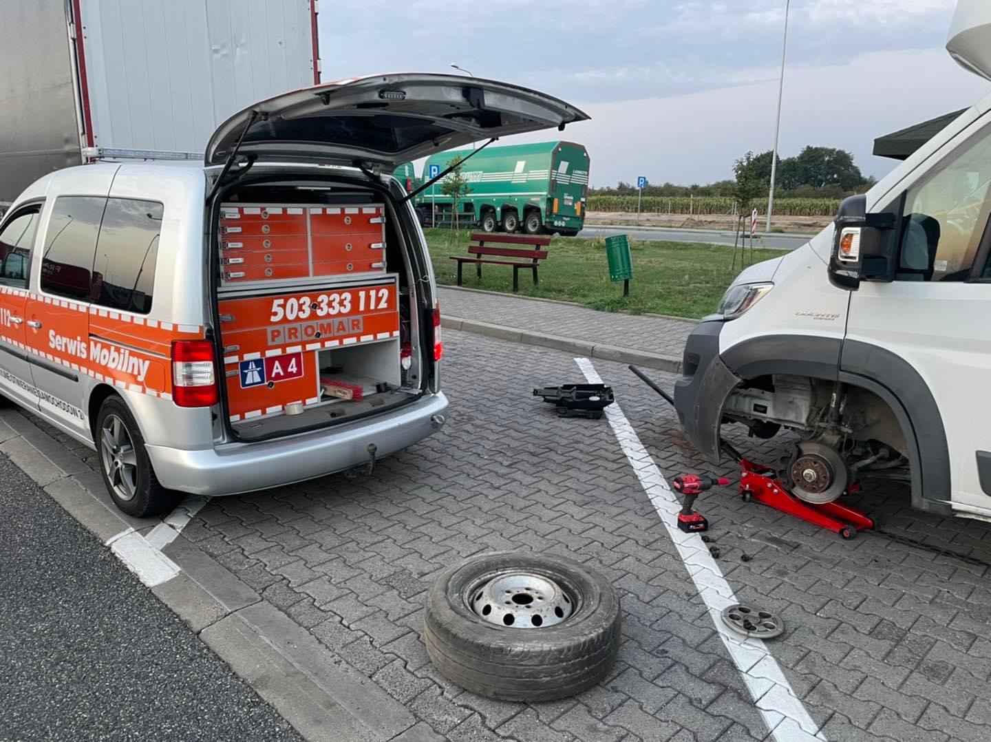 Mobilny serwis wymienia koło w dostawczym samochodzie na parkingu, widoczny bus serwisowy z otwartym bagażnikiem i narzędziami.