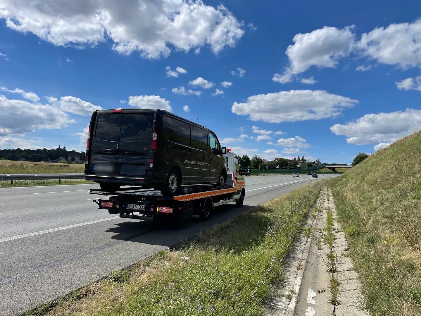 Czarny Opel Vivaro na lawecie pomocy drogowej, widok z tyłu na autostradzie w słoneczny dzień.