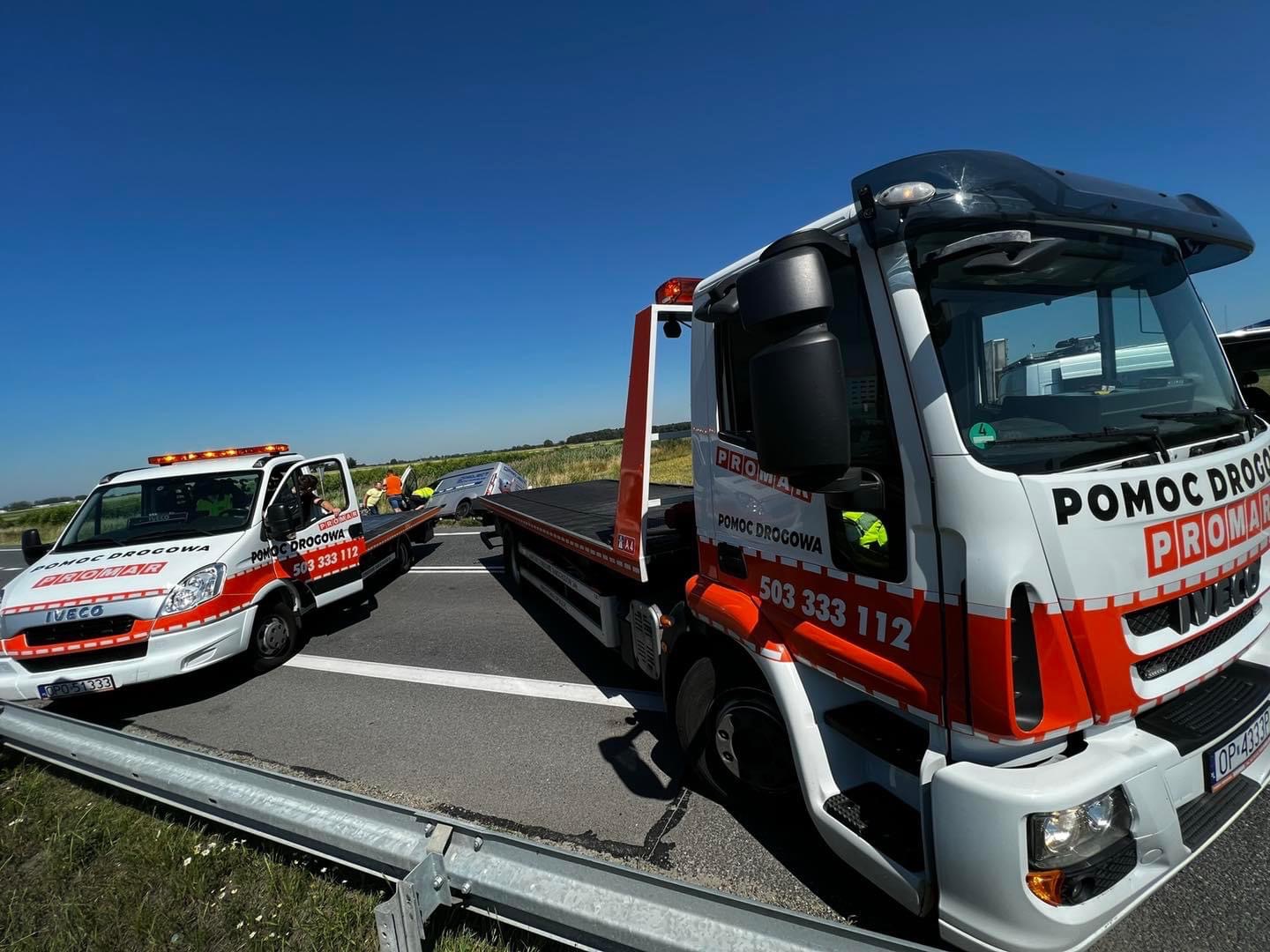 Dwa białe samochody pomocy drogowej marki Iveco z pomarańczowymi pasami i napisem 'POMOC DROGOWA PROMAR' na poboczu autostrady, jeden z lawetą, drugi z podnośnikiem, w tle uszkodzony samochód...