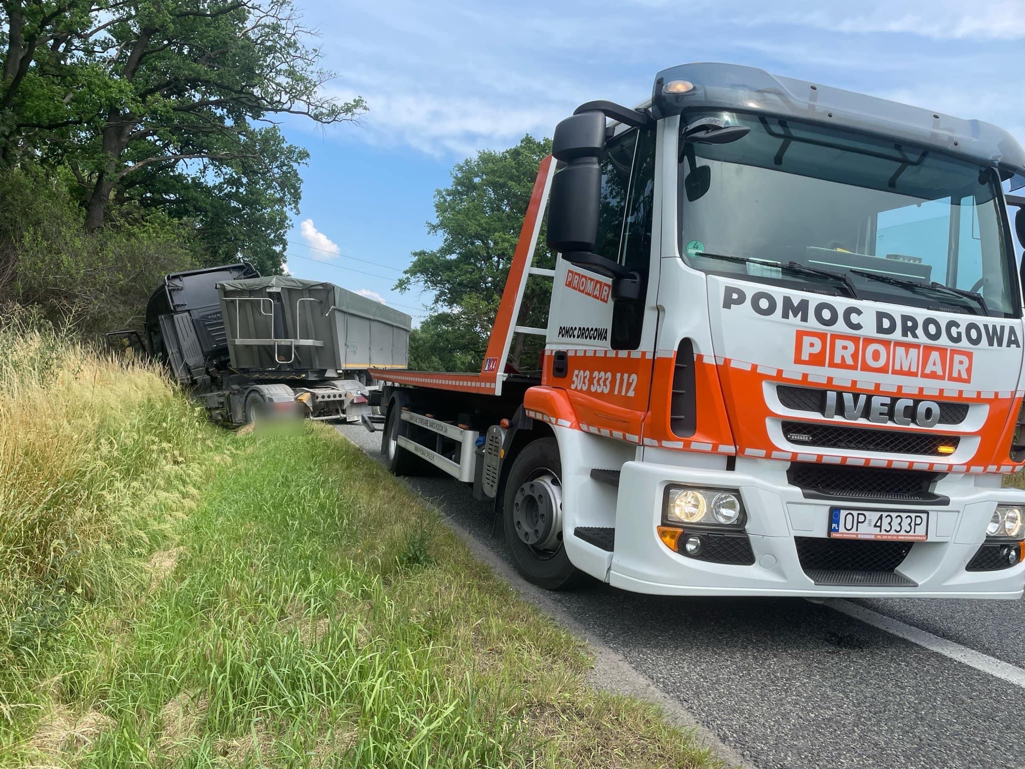 Ciężarówka pomocy drogowej marki Iveco, z pomarańczowymi elementami, holuje uszkodzoną ciężarówkę na poboczu drogi.