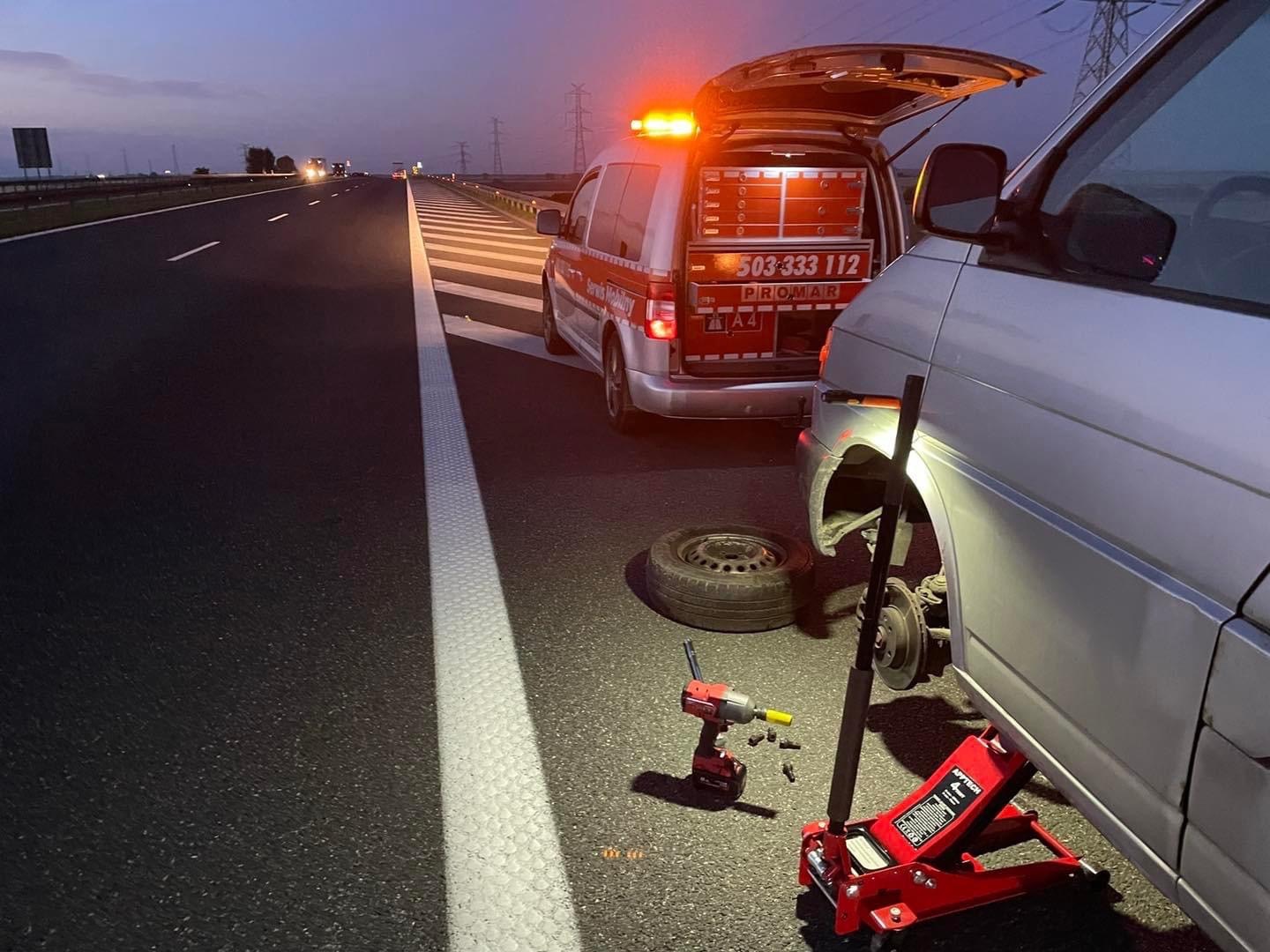 Wymiana koła w samochodzie osobowym na poboczu autostrady, widoczny podnośnik hydrauliczny, klucz udarowy i pojazd pomocy drogowej z otwartym bagażnikiem i światłem ostrzegawczym.