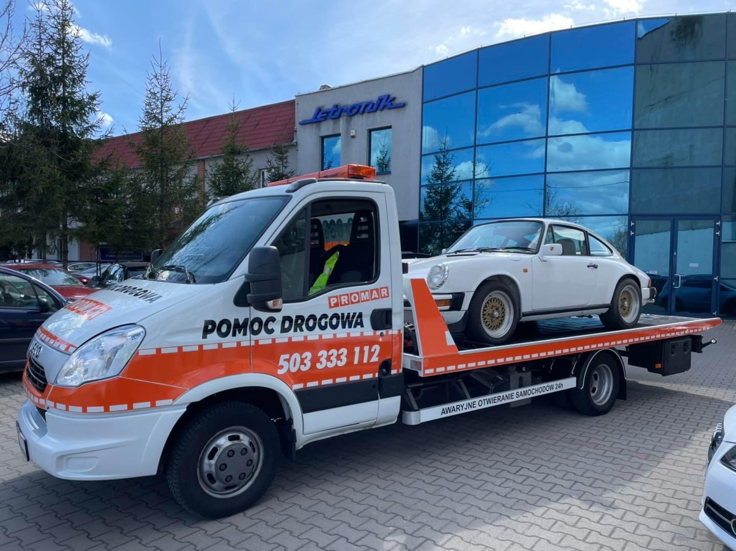 Biała laweta z pomarańczowymi elementami i napisem 'Pomoc Drogowa' przewozi białe sportowe Porsche na tle nowoczesnego budynku z niebieskimi szybami.