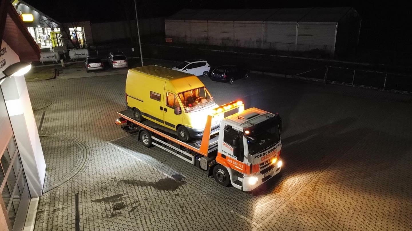 Żółty uszkodzony van na lawecie pomocy drogowej na parkingu w nocy, widoczny budynek stacji benzynowej w tle.