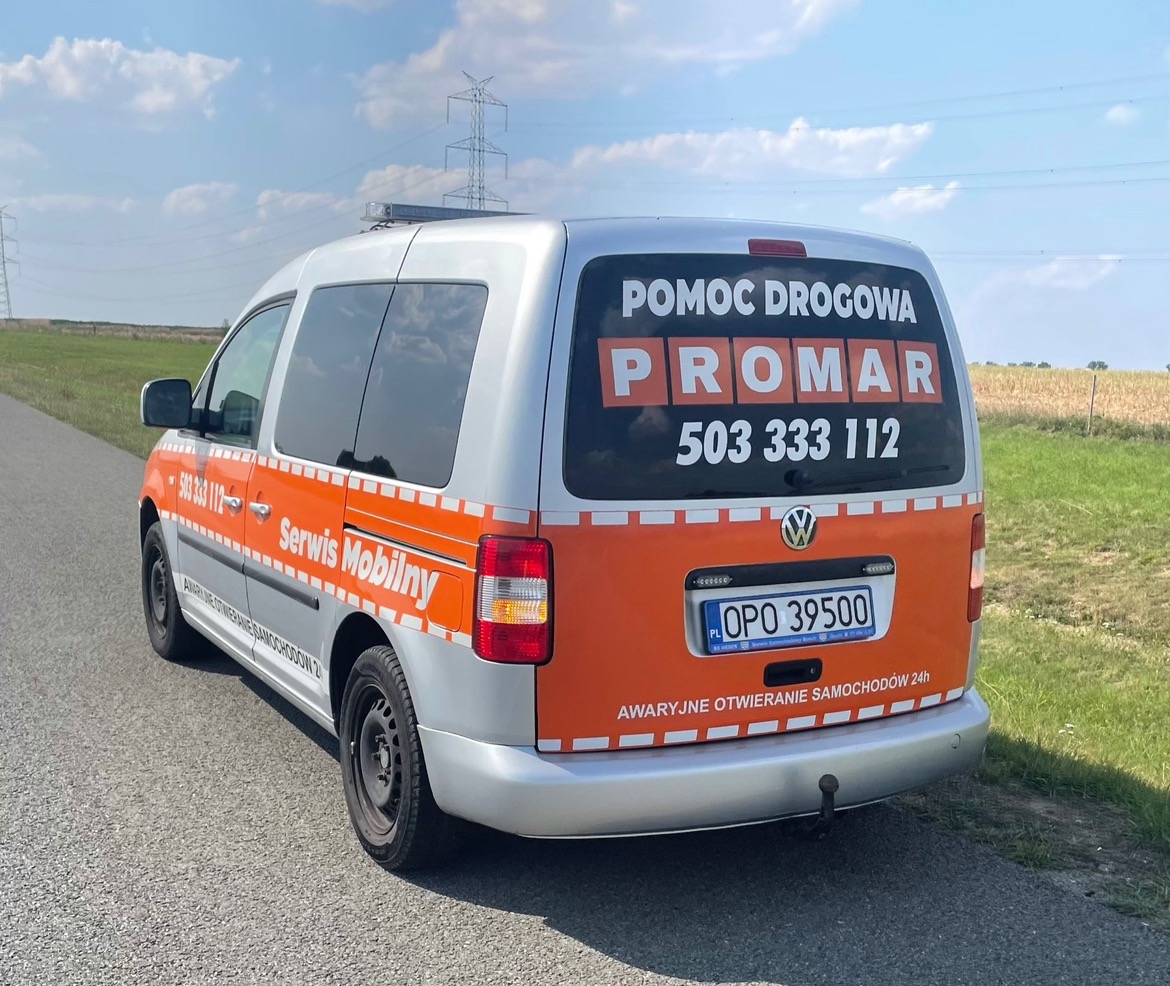 Srebrno-pomarańczowy samochód Volkswagen Caddy z napisem 'Pomoc Drogowa PROMAR 503 333 112' na tylnej szybie, stojący na poboczu drogi, z widocznymi słupami wysokiego napięcia w tle.