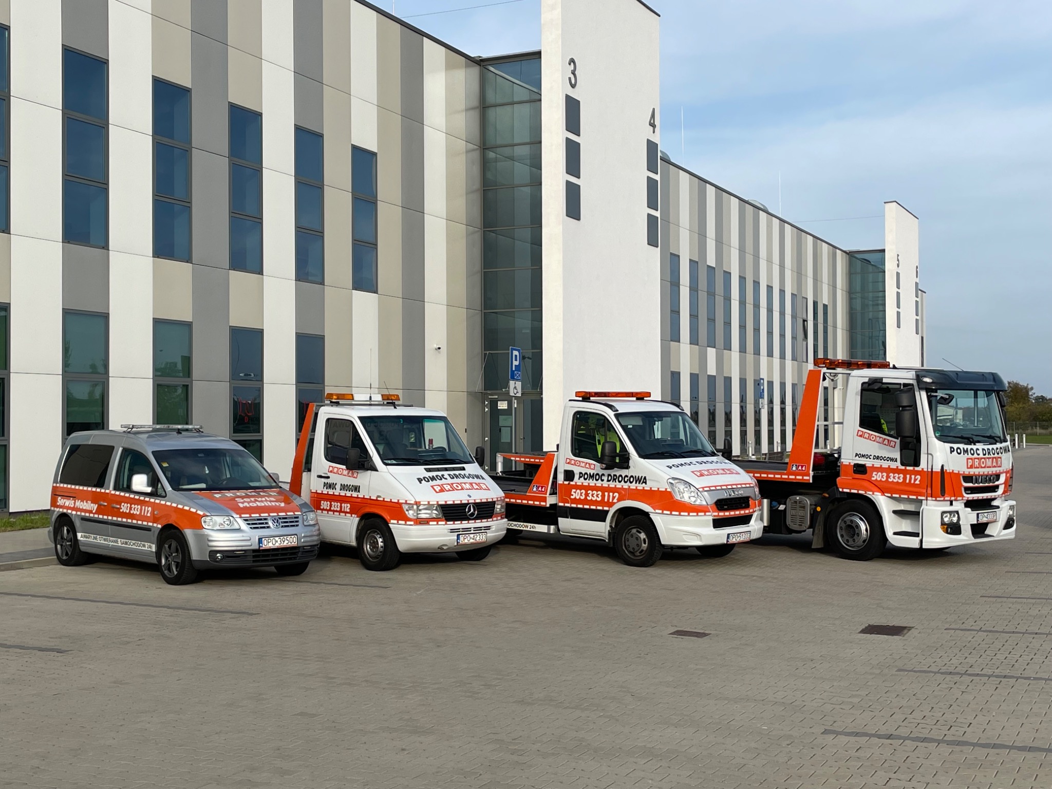 Flota pojazdów pomocy drogowej firmy Promar ustawiona przed nowoczesnym budynkiem biurowym, obejmująca od lewej: serwis mobilny Volkswagen Touran, dwa Mercedesy Sprintery oraz Iveco Eurocargo...
