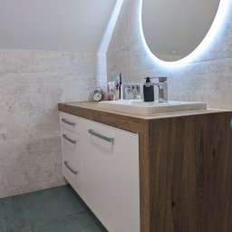 AT HOME - Nowoczesna szafka łazienkowa na wymiar z białymi frontami i blatem imitującym drewno, okrągłe lustro z podświetleniem LED, szarymi płytkami na ścianie i turkusową podłogą.