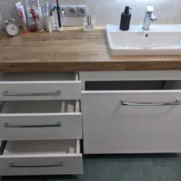 Biała szafka łazienkowa z drewnianym blatem, umywalką i otwartymi szufladami, widoczny zegar, kosmetyki i akcesoria do higieny osobistej.