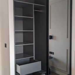 AT HOME - Szara szafa wnękowa z przesuwnymi drzwiami, widoczne półki i wysunięte dwie szuflady, minimalistyczny design z czarnymi ramami.