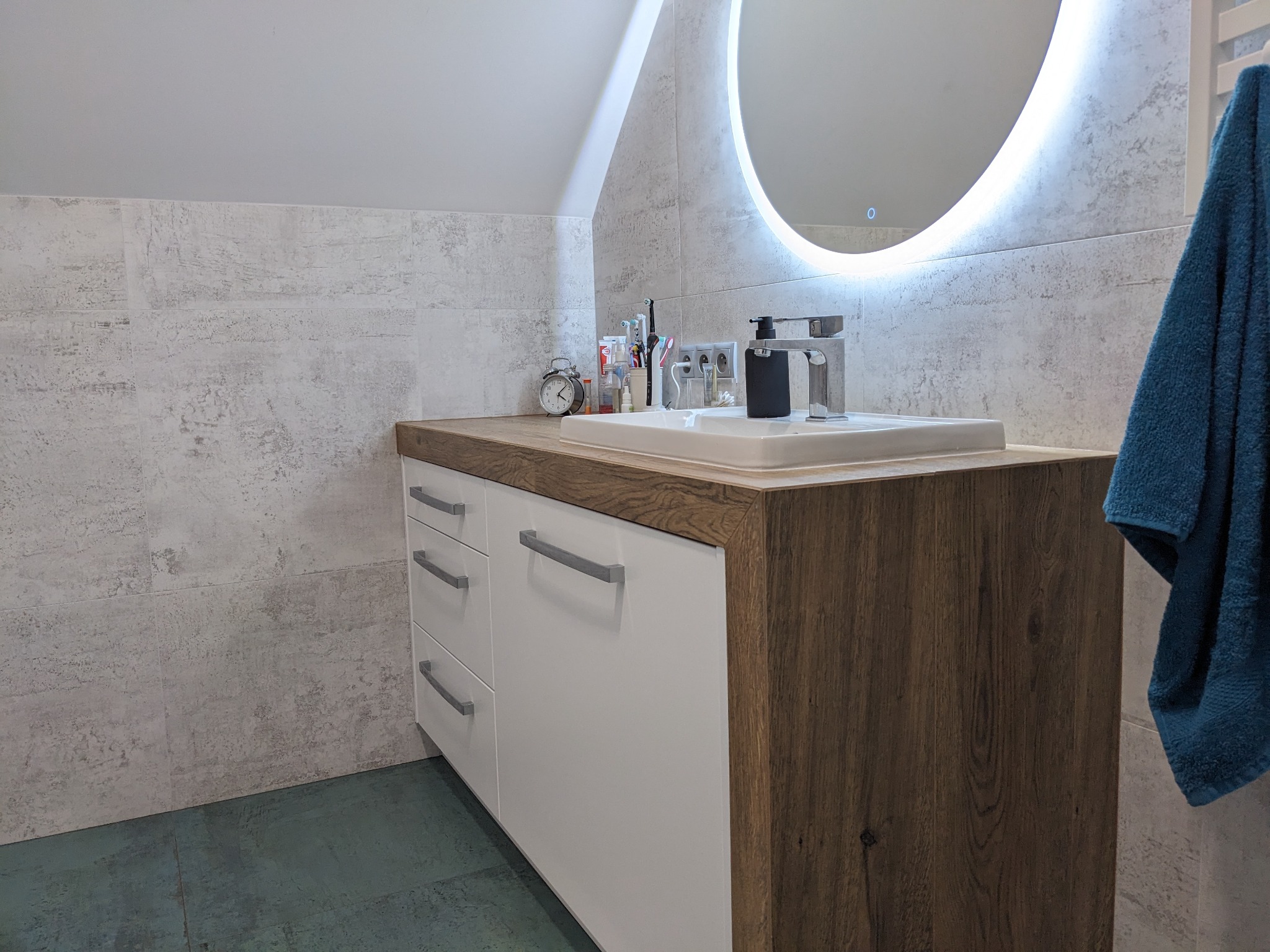 Nowoczesna szafka łazienkowa na wymiar z białymi frontami i blatem imitującym drewno, okrągłe lustro z podświetleniem LED, szarymi płytkami na ścianie i turkusową podłogą.