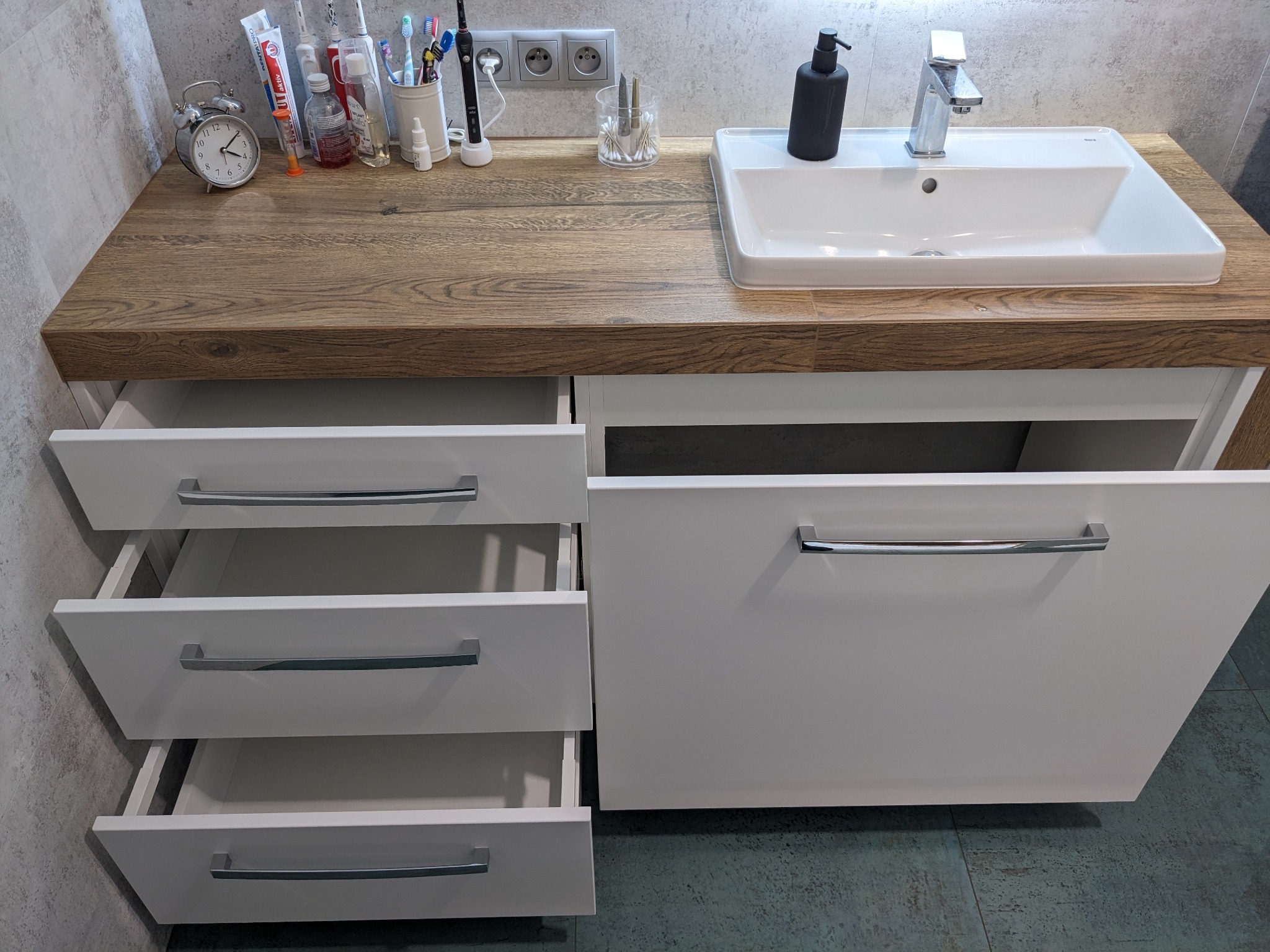 Biała szafka łazienkowa z drewnianym blatem, umywalką i otwartymi szufladami, widoczny zegar, kosmetyki i akcesoria do higieny osobistej.