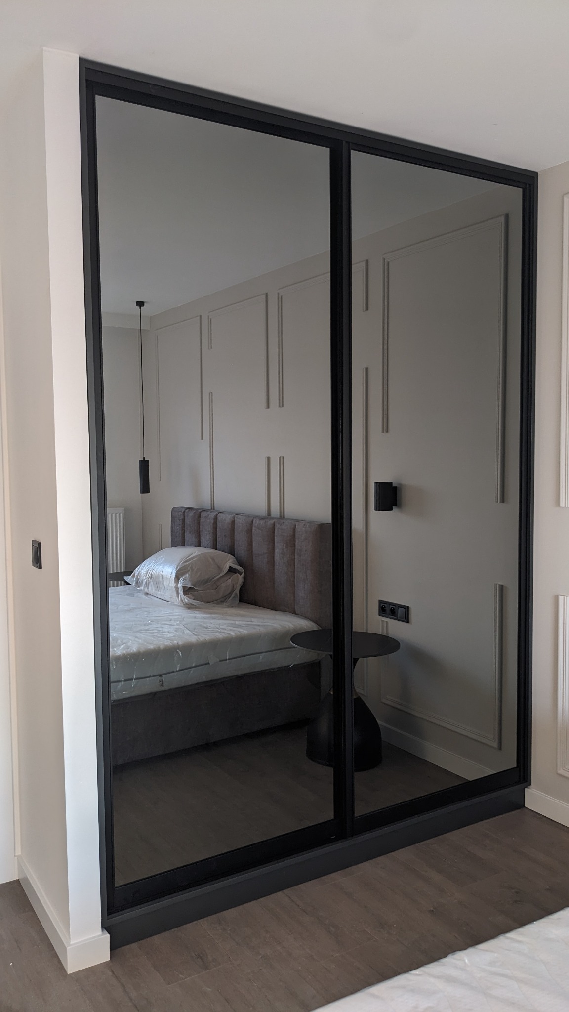 Szafa wnękowa z przesuwnymi drzwiami lustrzanymi w czarnej ramie, odbijająca wnętrze sypialni z łóżkiem i dekoracyjną ścianą.