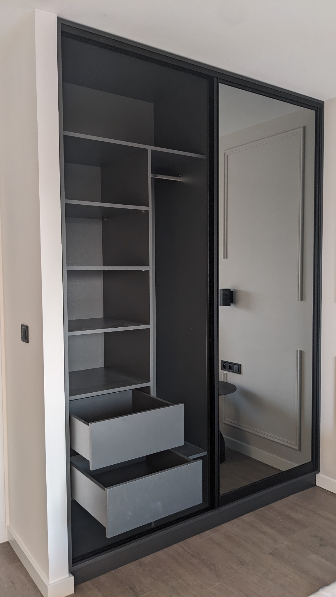 Szara szafa wnękowa z przesuwnymi drzwiami, widoczne półki i wysunięte dwie szuflady, minimalistyczny design z czarnymi ramami.