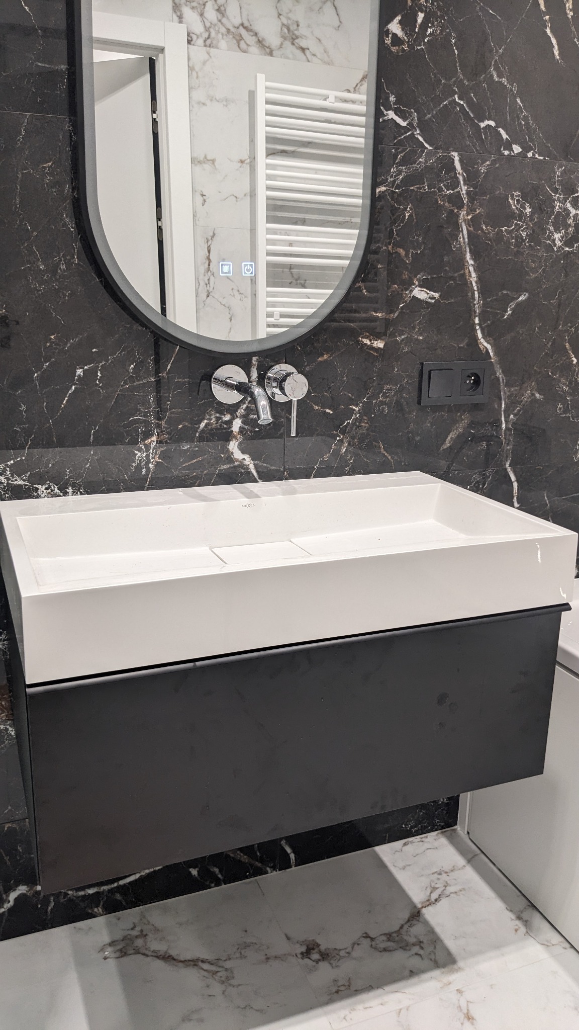 Nowoczesna łazienka z umywalką nablatową, czarnym blatem szafki i lustrem z podświetleniem na tle ściany z czarnego marmuru.