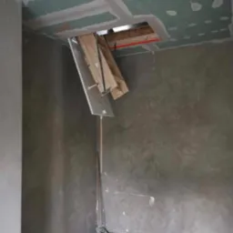 Montaż składanych schodów strychowych w otworze w suficie z płyt gipsowo-kartonowych, ściany w szarym tynku dekoracyjnym, widoczna szczotka do sprzątania.