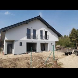 Usługi ogólnobudowlane - Dwukondygnacyjny dom jednorodzinny z szarą elewacją i czarnym dachem, w trakcie prac wykończeniowych na zewnątrz, widoczny przyczepka i materiały budowlane.