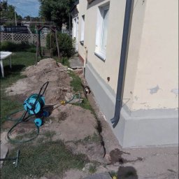 Usługi ogólnobudowlane - Ocieplenie fundamentów budynku jednorodzinnego, widoczny fragment ocieplonej ściany z szarą izolacją, obok leży zwinięty wąż ogrodowy, a przy ścianie siedzi mały kot.