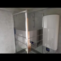 Nowoczesna kabina prysznicowa ze srebrnymi profilami i przezroczystym szkłem, obok biały, pionowy podgrzewacz wody z niebieskim panelem sterowania, na tle szare płytki ceramiczne.