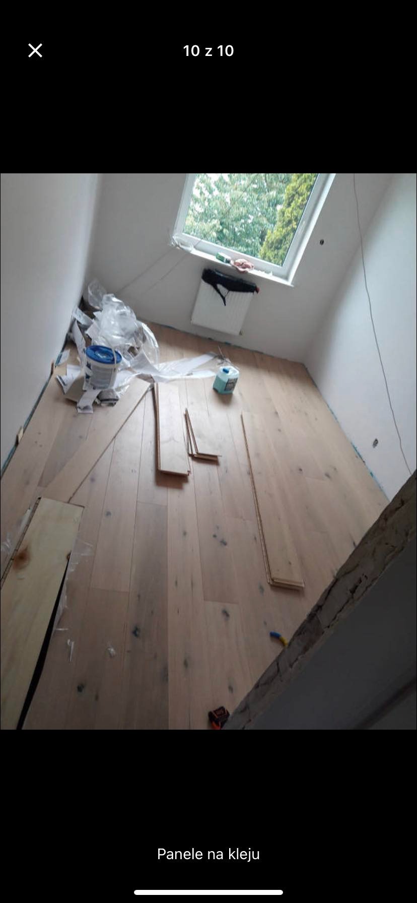 Montaż jasnych paneli podłogowych w pokoju na poddaszu z oknem dachowym, widoczne narzędzia, klej i resztki folii zabezpieczającej.