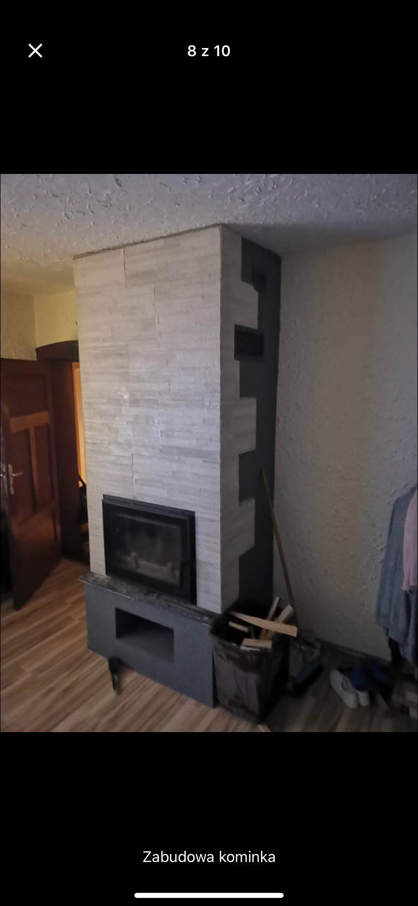 Obudowa kominka wykończona jasnymi płytkami imitującymi kamień, z czarnymi elementami po bokach, widok na wkład kominkowy i kosz z drewnem obok.