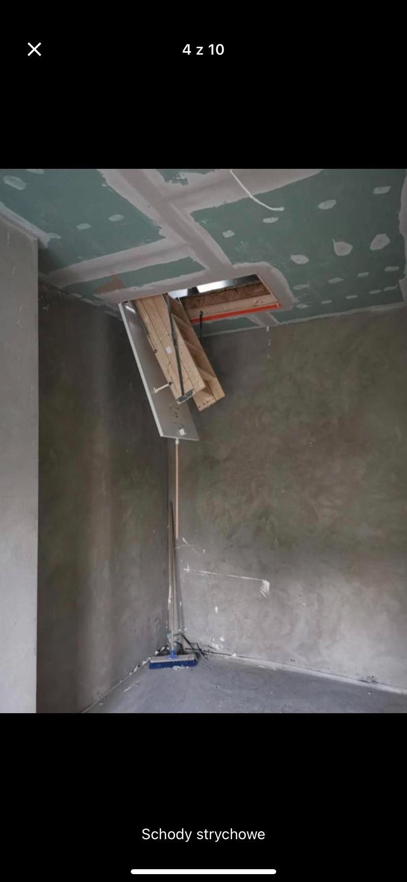 Montaż składanych schodów strychowych w otworze w suficie z płyt gipsowo-kartonowych, ściany w szarym tynku dekoracyjnym, widoczna szczotka do sprzątania.