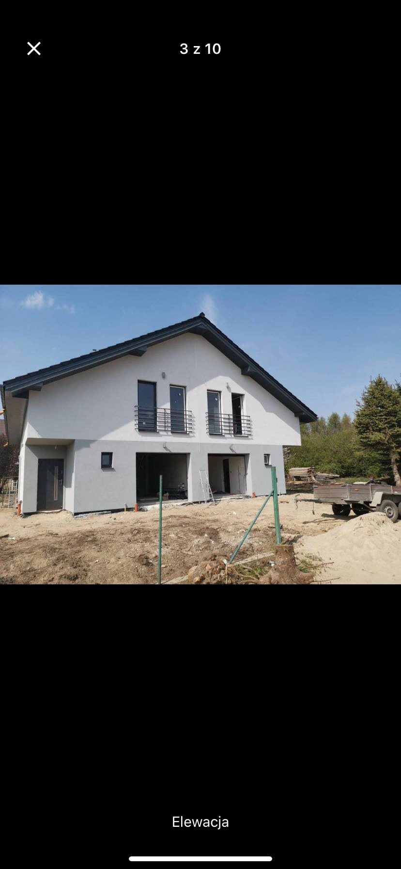 Dwukondygnacyjny dom jednorodzinny z szarą elewacją i czarnym dachem, w trakcie prac wykończeniowych na zewnątrz, widoczny przyczepka i materiały budowlane.