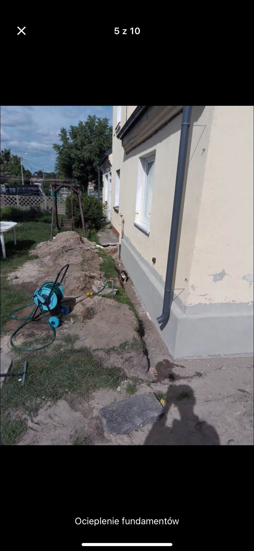 Ocieplenie fundamentów budynku jednorodzinnego, widoczny fragment ocieplonej ściany z szarą izolacją, obok leży zwinięty wąż ogrodowy, a przy ścianie siedzi mały kot.