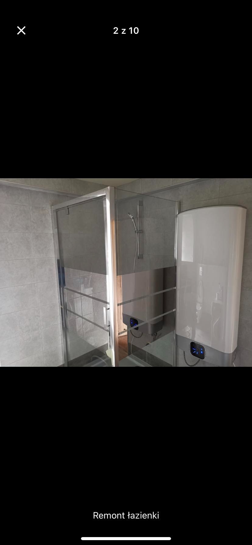 Nowoczesna kabina prysznicowa ze srebrnymi profilami i przezroczystym szkłem, obok biały, pionowy podgrzewacz wody z niebieskim panelem sterowania, na tle szare płytki ceramiczne.