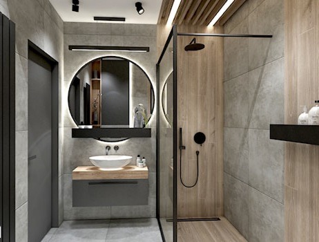 Nowoczesna łazienka z okrągłym lustrem podświetlanym LED, umywalką nablatową na drewnianym blacie, szarą szafką pod umywalką i kabiną prysznicową typu walk-in z czarną armaturą.