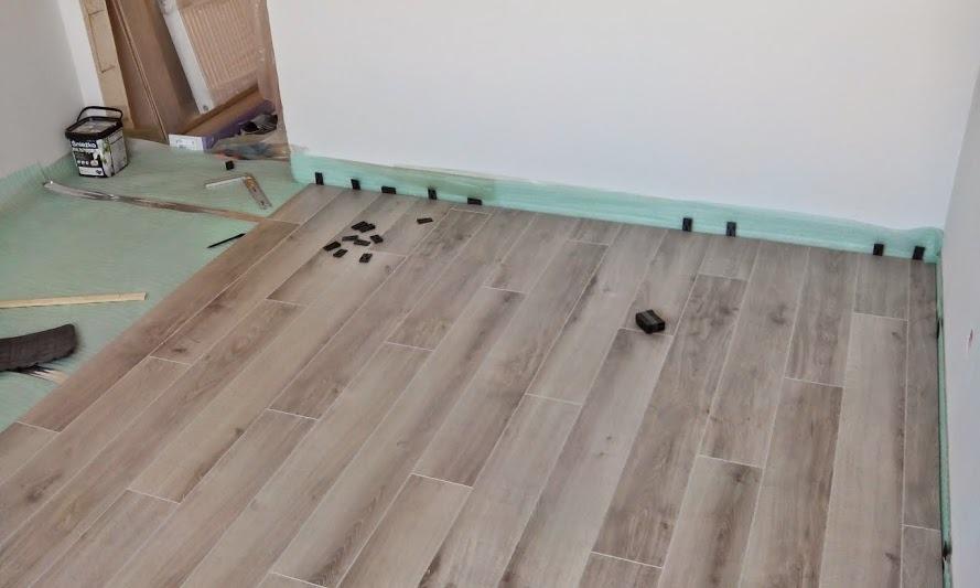 Układanie podłogi z płytek imitujących drewno, z wykorzystaniem klipsów dystansowych przy ścianie i narzędzi pomiarowych. Widoczna folia ochronna i wiaderko z klejem.