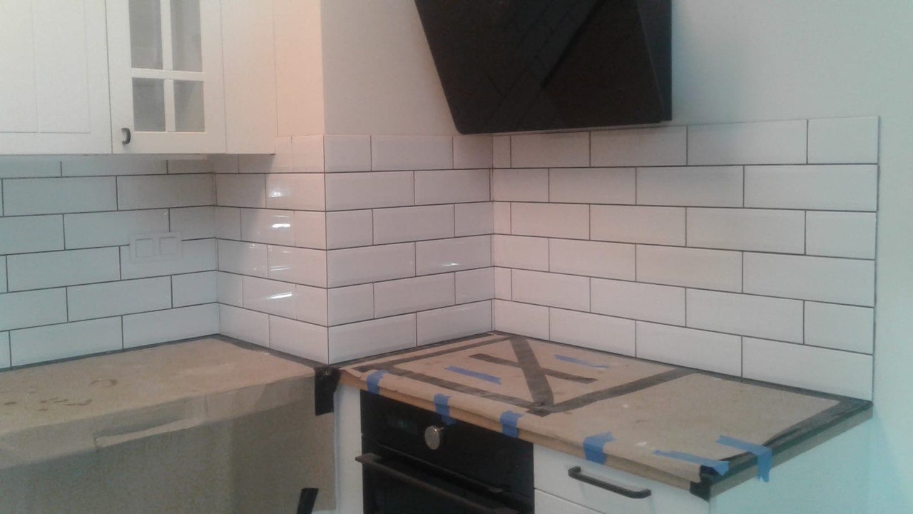 Kuchnia w trakcie wykończenia: białe, prostokątne płytki ułożone na ścianie nad blatem, zabezpieczony blat i sprzęty kuchenne folią ochronną i niebieską taśmą. Widoczny fragment szafki wiszącej.