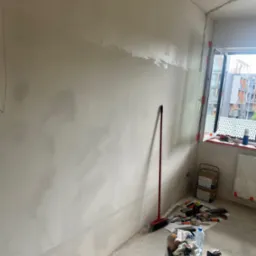 Ściana w trakcie szpachlowania, widoczne narzędzia, worki z zaprawą, oraz czerwona zmiotka oparta o ścianę, okno z widokiem na budynki w tle.