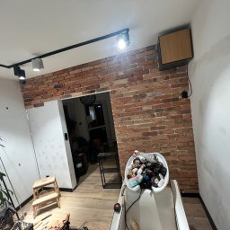 Laparis - Wnętrze w trakcie prac wykończeniowych: fragment ściany z cegły, sufit z oświetleniem szynowym, ślady po szpachlowaniu na ścianie, drabinka i akcesoria malarskie w pomieszczeniu.