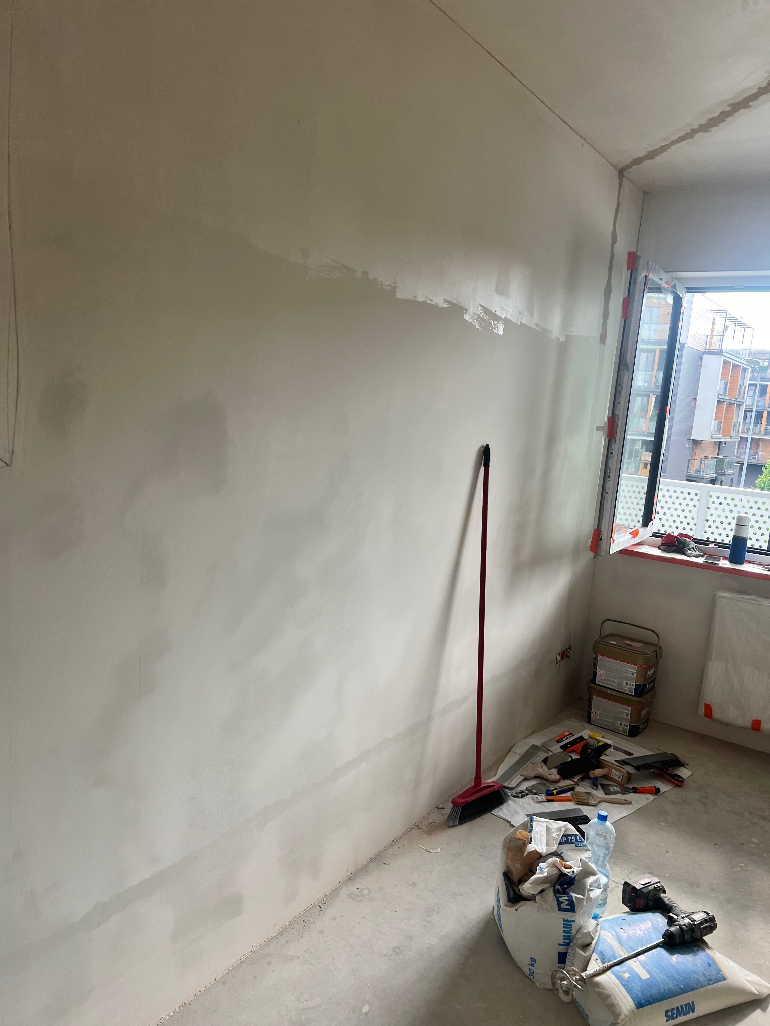 Ściana w trakcie szpachlowania, widoczne narzędzia, worki z zaprawą, oraz czerwona zmiotka oparta o ścianę, okno z widokiem na budynki w tle.