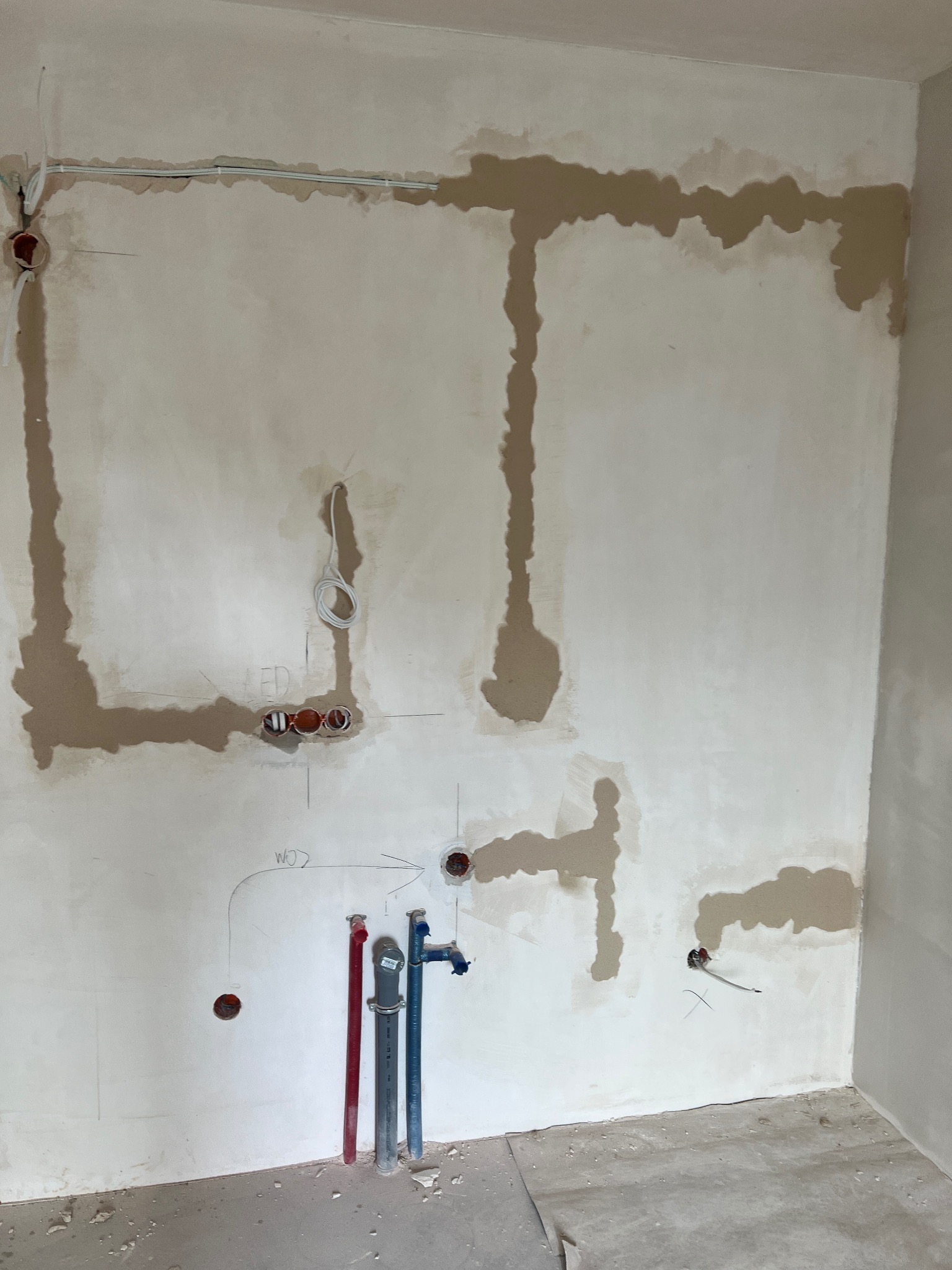 Ściana w trakcie remontu z widocznymi śladami po instalacjach elektrycznych i hydraulicznych, otwory w ścianie, rury wystające z podłogi, częściowo zaszpachlowane bruzdy. Na ścianie widoczne...