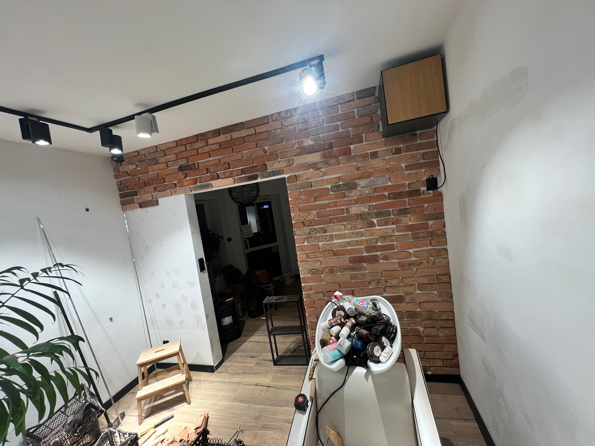 Wnętrze w trakcie prac wykończeniowych: fragment ściany z cegły, sufit z oświetleniem szynowym, ślady po szpachlowaniu na ścianie, drabinka i akcesoria malarskie w pomieszczeniu.