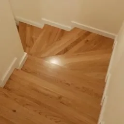 Widok z g&oacute;ry na drewniane schody z jasnego drewna, z białymi listwami przypodłogowymi i ścianami, ukazujące bieg schod&oacute;w skręcający w prawo.