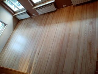 Jasny, odnowiony parkiet z wyraźnym usłojeniem, widziany z góry, z fragmentem schodów po prawej stronie i grzejnikami pod oknem.