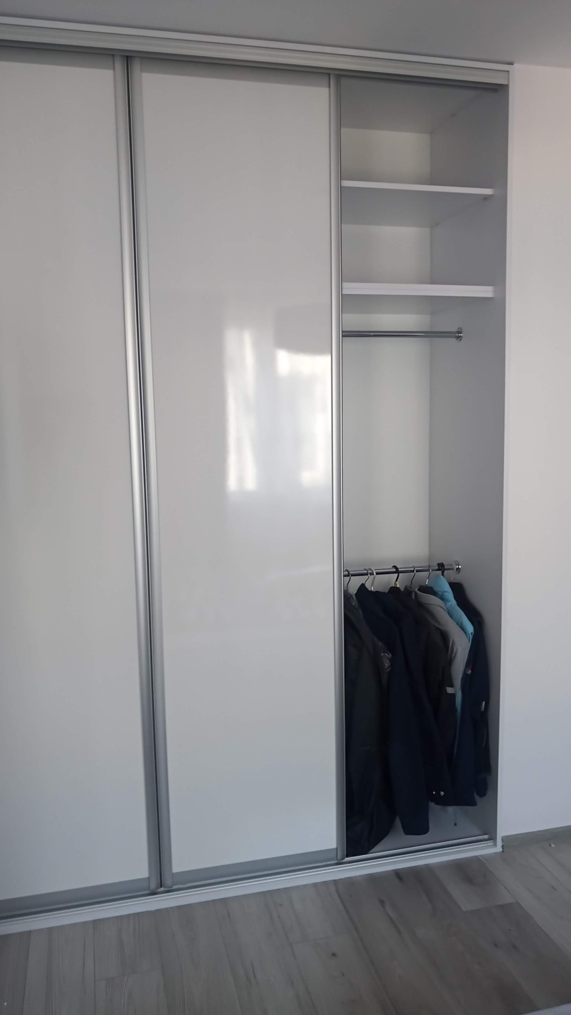 Biała szafa przesuwna wnękowa z częściowo otwartymi drzwiami, widoczne półki i wiszące ubrania.
