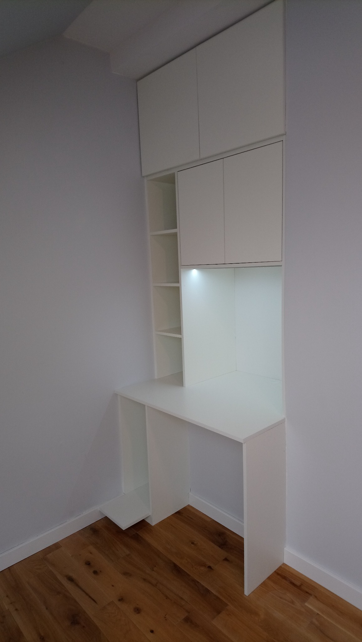 Białe biurko na wymiar z szafkami wiszącymi i półkami, oświetlenie LED pod szafkami, drewniana podłoga, minimalistyczny design.