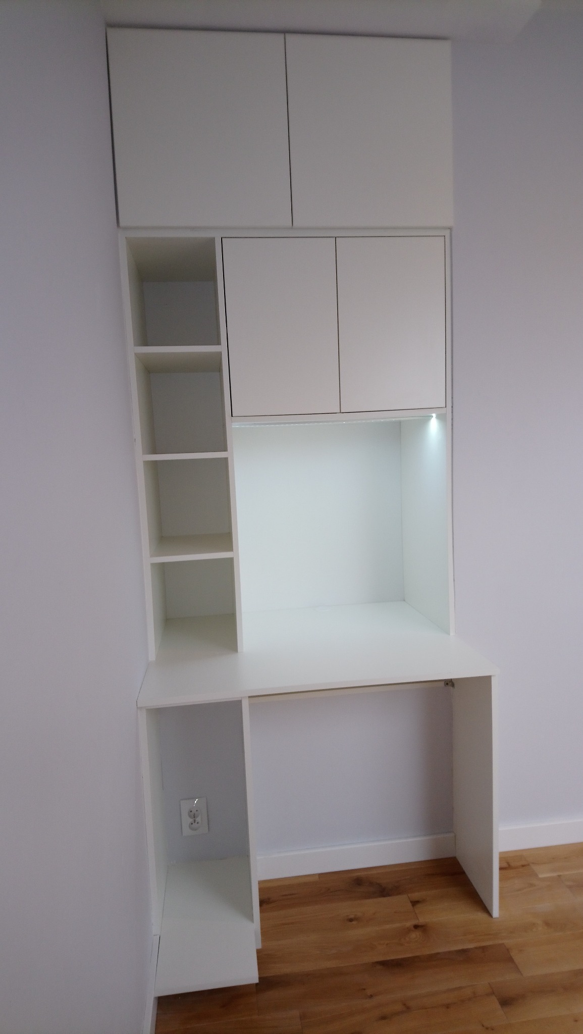 Białe biurko na wymiar z nadstawką szafek i półkami, oświetlenie LED pod szafkami, widoczny kontakt w dolnej części biurka.