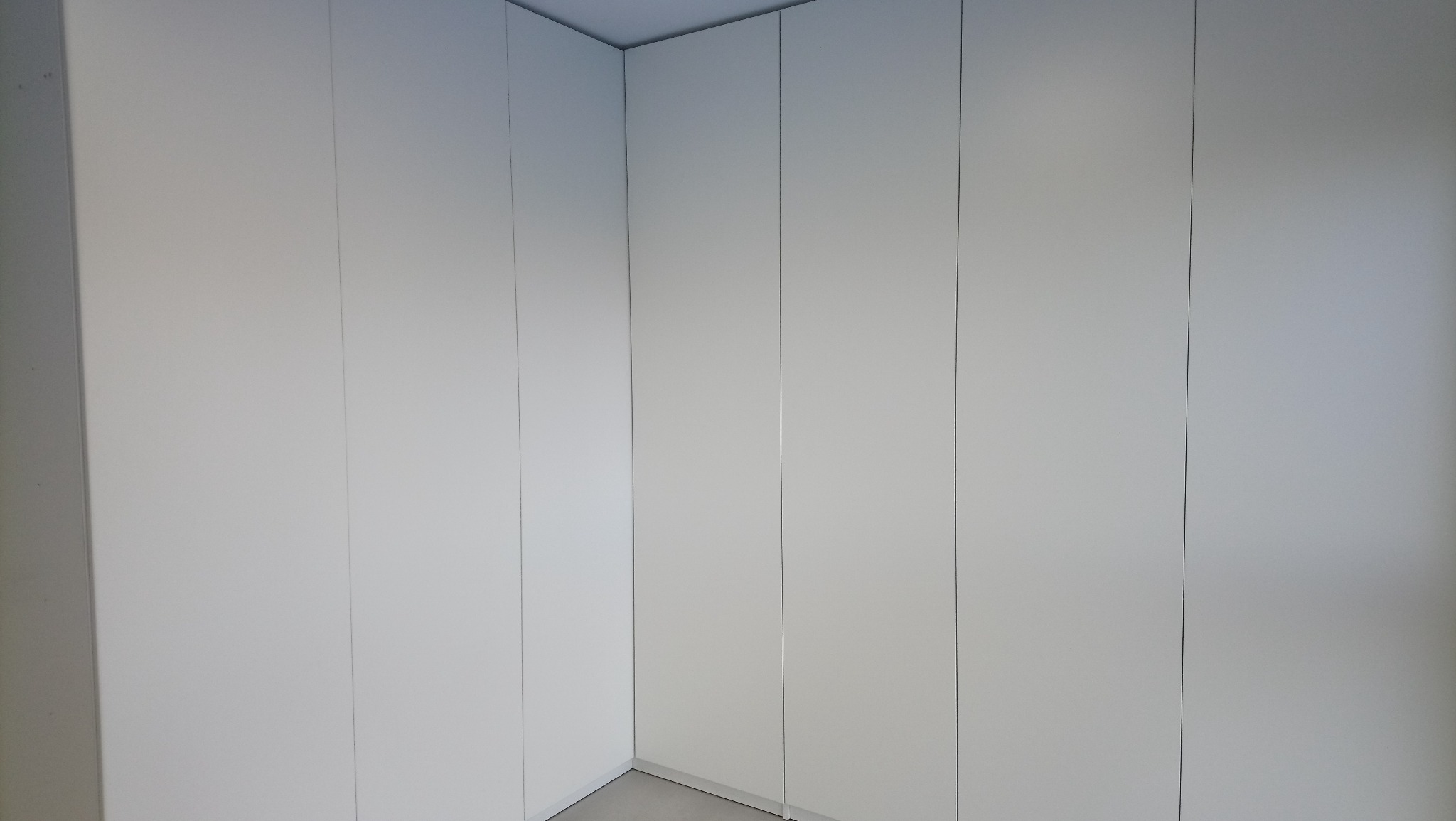 Minimalistyczna, biała szafa wnękowa z gładkimi frontami, wypełniająca przestrzeń od podłogi do sufitu, widoczna z narożnika pokoju.