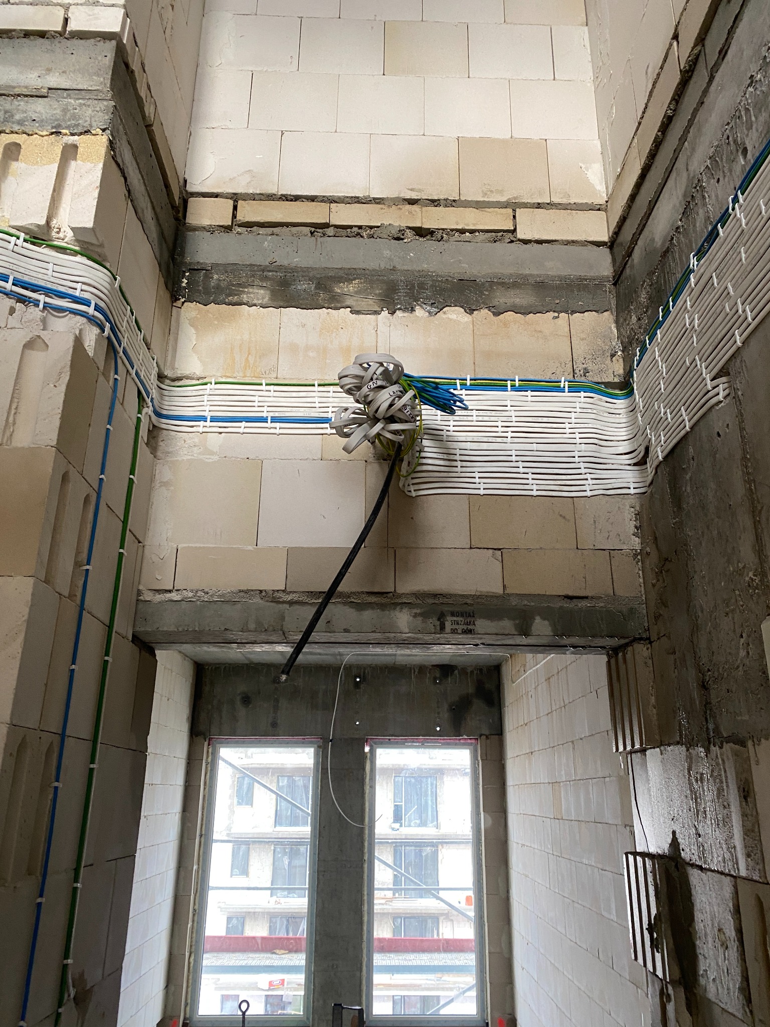 Wewnętrzne ściany budynku w trakcie budowy z widocznymi przewodami elektrycznymi prowadzonymi w korytach kablowych, ze zwojem kabli wychodzącym z otworu w ścianie i oknem w tle.