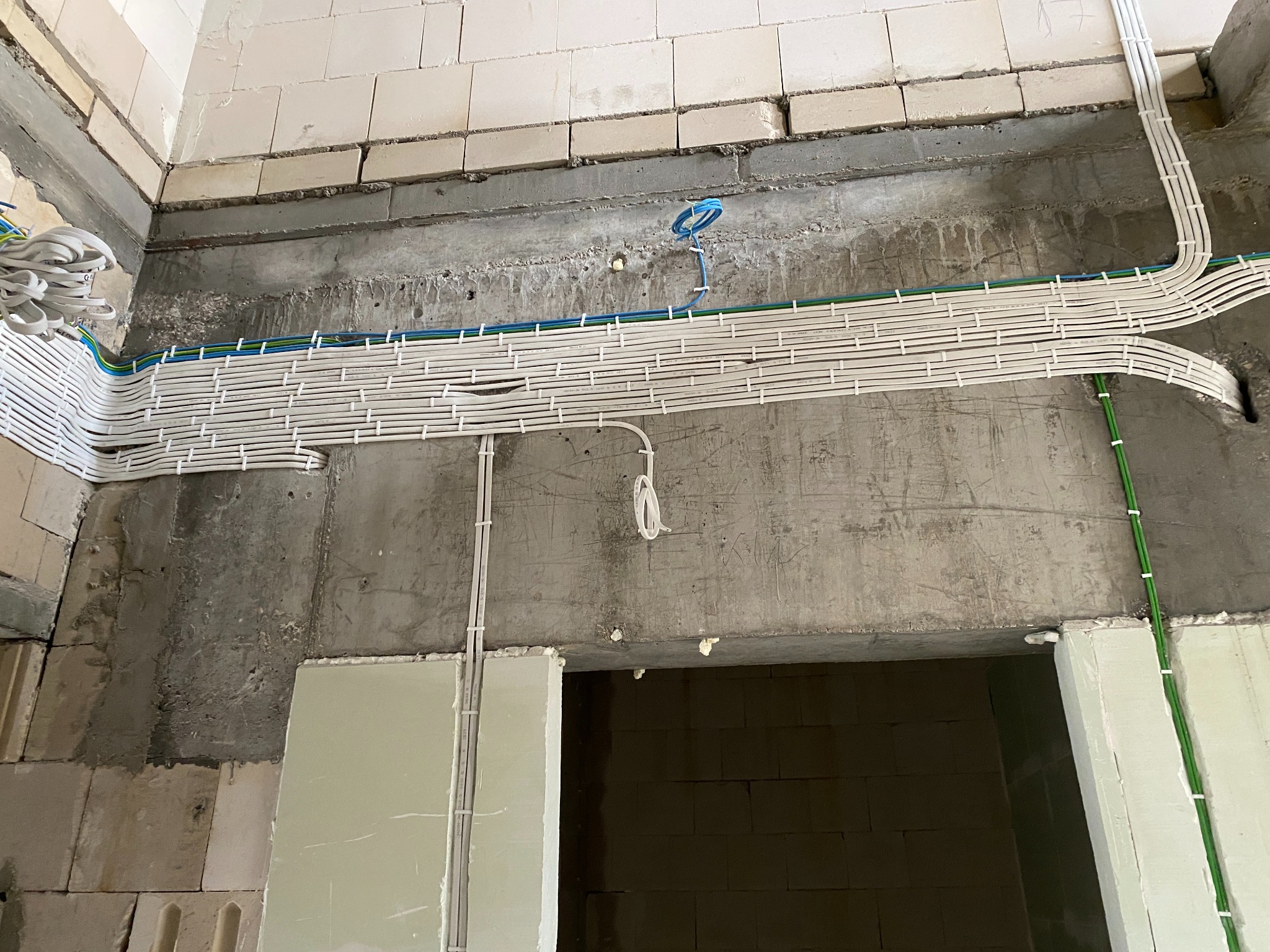 Równoległe, białe kable elektryczne ułożone w plastikowych korytkach na surowej, betonowej ścianie w trakcie remontu. Widoczne również kable w peszlach. Część przewodów zwisa luźno.