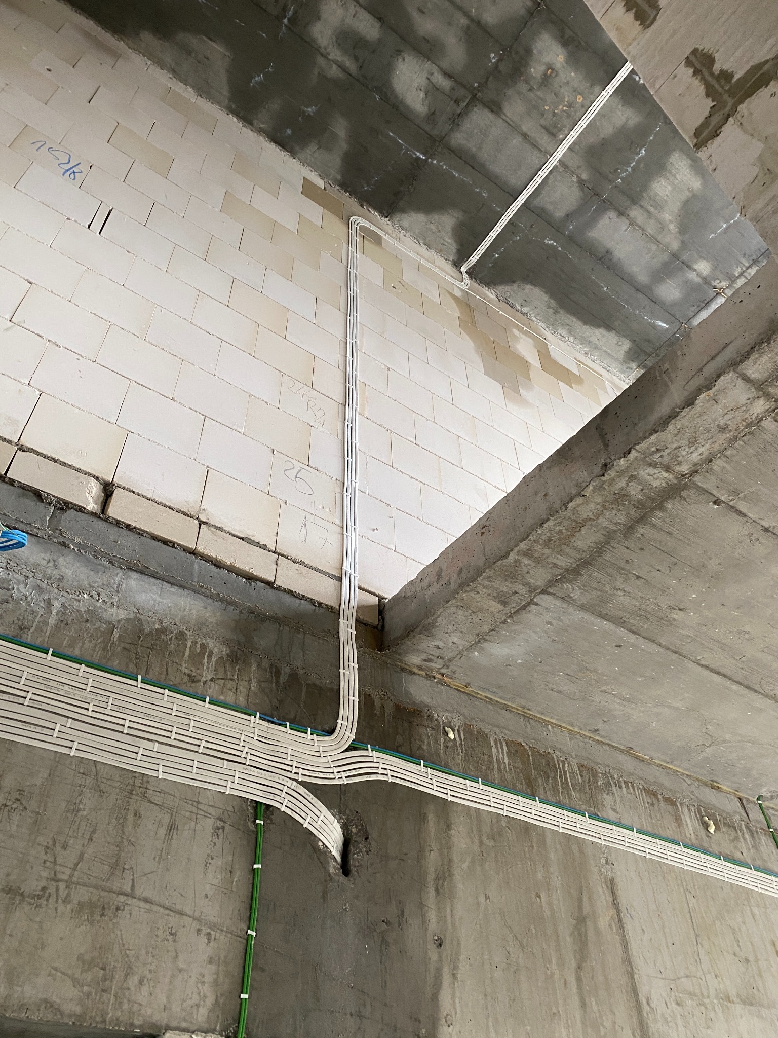 Białe kable elektryczne ułożone w korytkach na surowych ścianach i suficie w trakcie instalacji w budynku w stanie deweloperskim, widoczne zielone peszle zabezpieczające kable.